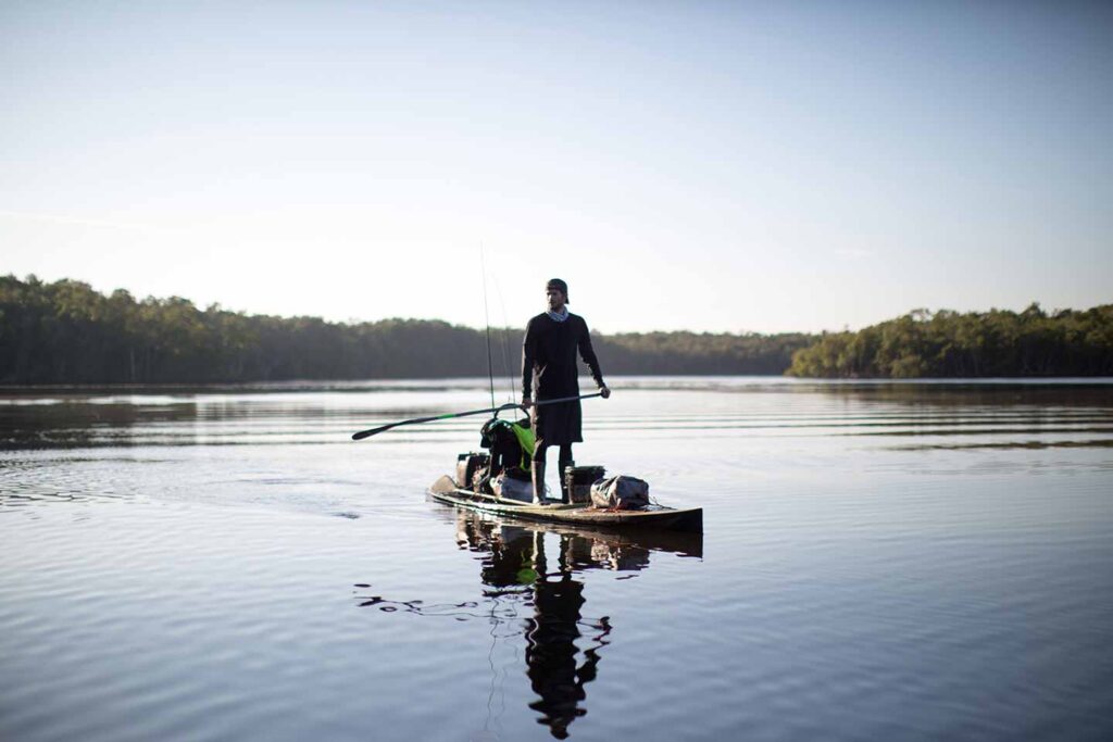 Person fishing from a paddleboard on calm water, surrounded by scenic nature