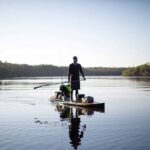 Person fishing from a paddleboard on calm water, surrounded by scenic nature